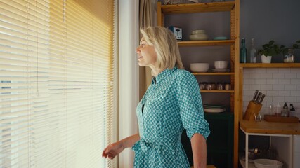 Happy retired woman smiling and enjoying her free time, standing in a modern kitchen next to a window with blinds, expressing serenity and positivity - Powered by Adobe