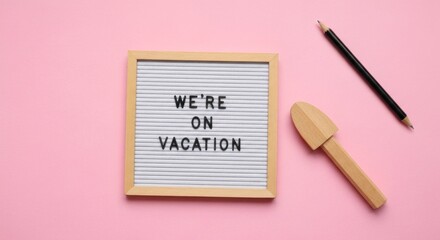 A small, light-wooden letter board displays "We're On Vacation". A small wooden tool and pencil rest beside it on a pink surface