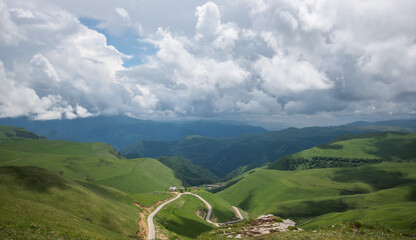 Sweeping view of a verdant valley with a winding road leading to a small settlement, framed by rolling green hills and a dramatic sky with dynamic cloud formations
