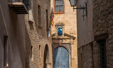 Fototapeta premium A narrow alley in a historic Spanish town, featuring stone buildings and a beautifully ornate doorway with flags and floral decorations. Old-world charm and cultural heritage in a picturesque setting