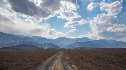 a landscape depicting a wide, dry grassland leading to a dirt road that stretches towards distant mountains under a partly cloudy sky