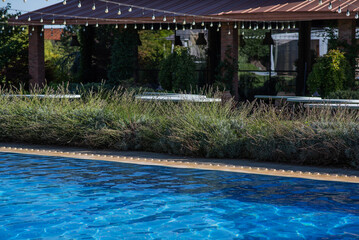 poolside relaxation area with a covered outdoor seating space, lush greenery, and string lights hanging above, featuring a blue water pool, wooden benches, and potted plants in a peaceful garden