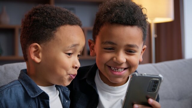 Two African American little boys children scrolling phone at home smiling satisfied enjoying smartphone mobile apps typing laughing friendship family brothers kids sons siblings with modern technology - Powered by Adobe
