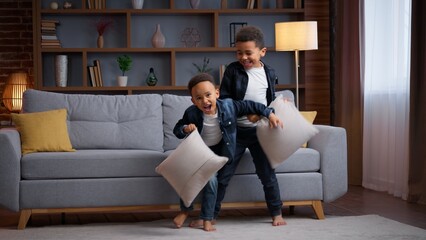 Two playful little African American kids fighting with pillows at home boys siblings brothers sons friends children enjoy fun playing funny fight battle game bit with cushions happiness family play