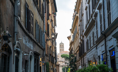 Narrow alley in central Rome with tall historic buildings, shuttered windows, and classic iron lanterns, leading to a distant church tower surrounded by terracotta rooftops and stone pine trees.