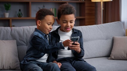 Two cute African American siblings boys with phone in living room using mobile cyberspace technology using smartphone together internet addicted sons children kids brothers watching online funny video