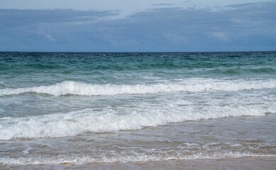 Foamy waves gently kiss the sandy shore, a dance of sea and land under the expansive blue sky, the essence of peaceful beach side tranquility