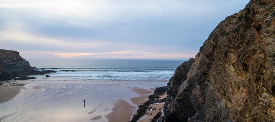 A peaceful coastal scene at sunset with a solitary figure walking on the sandy beach. The waves gently lap the shore, and towering cliffs frame the coast of Portugal.