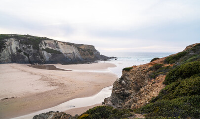 The rugged coastline of Alentejo in Portugal features dramatic cliffs meeting a secluded sandy beach. The tide has receded, revealing intricate patterns on the wet sand.