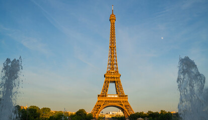 Eiffel Tower in Paris illuminated by soft evening light, framed by water fountains on either side, with the moon visible in the clear blue sky, creating a classic Parisian scene.