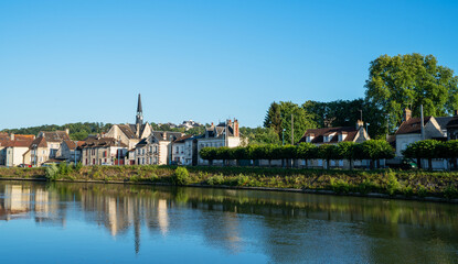 A picturesque French village nestled along a calm river, with reflections of charming houses and a church spire on the water. The scene is bathed in warm sunlight under a clear blue sky.
