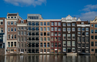 Row of colorful, narrow canal houses with large windows and gabled roofs reflecting on a calm waterway under a blue sky, located in a picturesque city center.