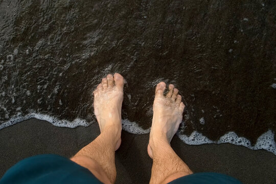 Gentle waves washing over barefooted man legs while standing on volcanic black sand beach in peaceful tropical vacation moment. Tourism travel journey trip. Wonderful wild nature dream place.