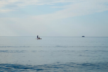 Couple man woman enjoying stand-up paddle boarding in calm sea waters. Romantic trip in dream place in evening. Blue seascape in soft colors with human silhouettes. Travel tourism vacations.