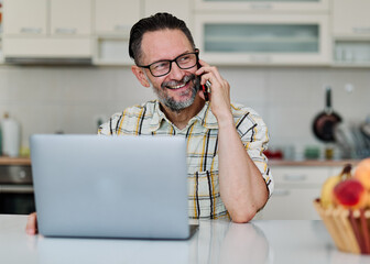 Portrait of a middle aged man or businessman working from home office using laptop and smart phone ...