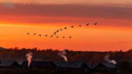 Geese flying in formation over a rural village at sunset with smoke rising from rooftops