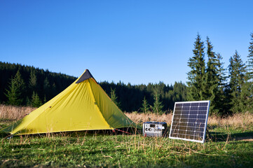 Tourist tent with solar panel and portable power station nearby in grassy field. Scene surrounded by rolling hills, tall pine trees under clear blue sky, showcasing eco-friendly camping setup. © anatoliy_gleb