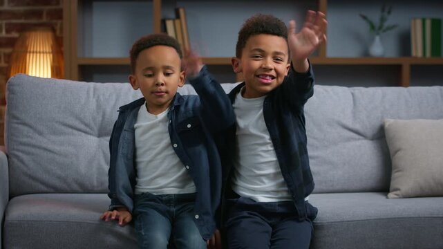 Two funny African American children greeting hello at home happy carefree boys kids siblings brothers sons in living room together looking at camera waving hands video call family joy fun childhood
