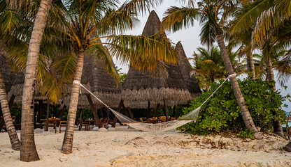 tropical beach with a white hammock tied between palm trees on soft sand, surrounded by lush greenery and traditional thatched-roof huts with wooden tables and chairs, located in a seaside resort
