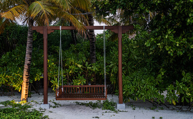 Fototapeta premium Wooden swing with rope hanging beneath palm trees on white sand in the Maldives, surrounded by dense tropical foliage and soft beach lighting, creating a peaceful coastal retreat in the evening light.