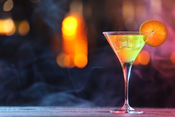 Glass of alcoholic cocktail with garnish on table against blurred background with smoke, space for text. Bokeh effect