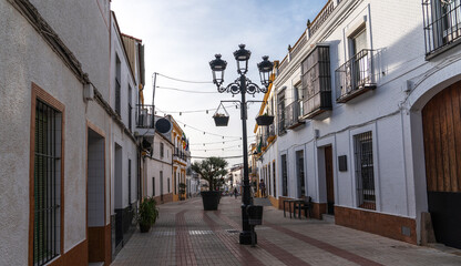 A peaceful pedestrian promenade stretches through a Spanish town, flanked by white houses and punctuated by large potted olive trees under a clear blue sky.