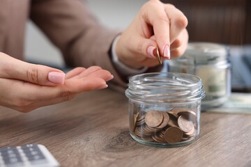 Woman putting coin into glass jar at wooden table indoors, closeup