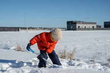 Child in winter clothes shoveling snow outdoors on a sunny day near suburban houses in cold weather