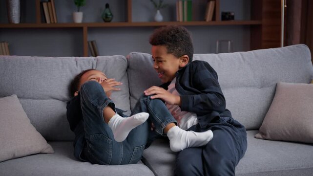 Two African American children tickling talking at home cute brothers little family kids boys sons siblings playing together having fun on couch laughing play fighting game fooling around childhood joy
