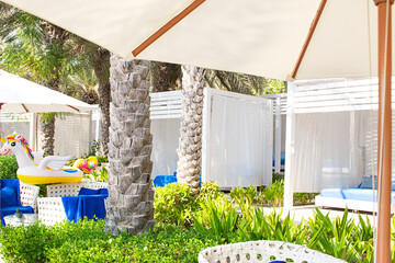 Luxury cabanas or tent with white curtains and blue lounge chairs under palm trees and umbrellas. Green plants and inflatable unicorn float create tropical summer resort atmosphere