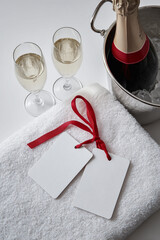 Two empty white tags tied with a red ribbon lie on a terry towel, next to two glasses of champagne and a bucket of ice with a bottle of champagne
