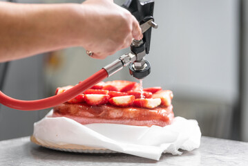 A pastry chef applying icing to a cake with a professional icing gun. The chef hands are clearly visible handling the tool, while the icing carefully covers the cake surface. The cake sits on