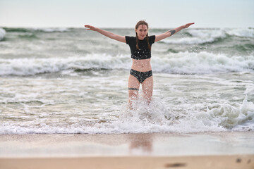 Young female with red hair performs yoga-like stance in baltic sea. Overcast sky and grey waves create serene, energetic atmosphere. Water splashes around her legs