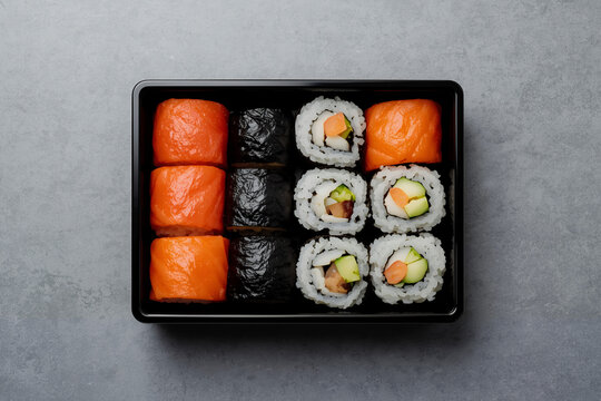 Top view sushi box with twelve maki rolls in black tray on gray, salmon avocado cucumber filling, clean minimal layout.