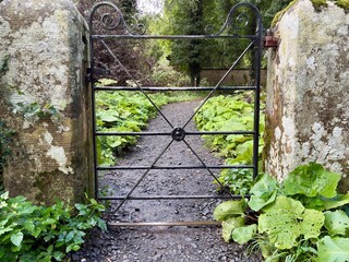 A quaint iron gate stands between two weathered stone pillars, leading to a lush garden path lined with greenery and framed by trees. A serene, rustic scene perfect for countryside charm.