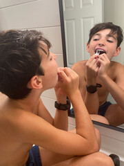 A young boy is meticulously cleaning the dental braces on his teeth in the bathroom mirror.
