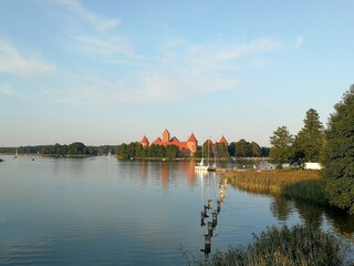 Panoramic view of Trakai Castle in Lithuania, a medieval red brick castle, situated on an island surrounded by a serene lake. The image captures the castle's towers and walls reflecting on the calm wa