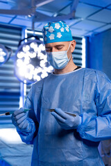 Surgeon in blue uniform holding surgical instruments. Surgeon in protective mask and blue uniform holding surgical instruments in operating room.