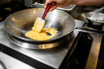 Chef cooking scrambled eggs in a wok on induction cooktop in professional kitchen. Early stage of cooking, stainless steel background, culinary process in action