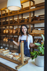 Smiling Woman Bakery Owner Behind Fresh Bread Display In Cozy Shop With Wooden Shelves And Warm Lighting