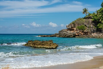 Fototapeta premium Turquoise ocean waves gently lap onto a sandy beach with a rocky cliffside and palm trees under a blue sky