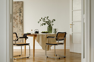 Dining setup with wooden table, woven chairs, eucalyptus in a vase and black teapot on linen cloth.