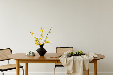 Dining table with yellow forsythia flowers in a dark vase, limes, and woven chairs in a bright interior.