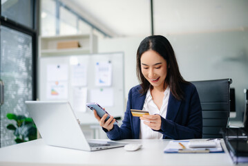 Confident Asian businesswoman smiling while using smartphone and credit card at office desk.