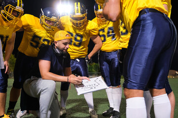 Football coach showing play strategy to American football players huddling on field at night