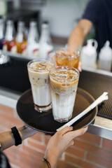 A barista's hands prepare 3 glasses of coffee milk on a tray to be served.
