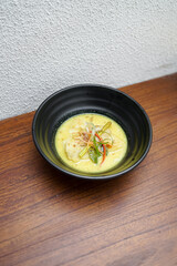 A black bowl filled with chicken in yellow curry sauce, topped with sliced vegetables and herbs. The dish looks creamy, spicy, and ready to serve for lunch or dinner .
