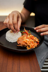A hand picks food from a plate of white rice and crispy chicken topped with spicy sambal and colorful vegetables. The meal is served fresh and hot, ideal for lunch or casual dining .