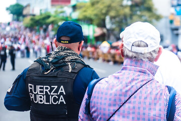 Public force officer and elderly man watching a parade on a city street, with crowd and colorful...