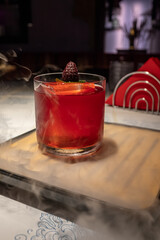 Smoky red cocktail served in a short glass with ice cube and berry garnish, presented on a wooden tray surrounded by dramatic smoke effect, ideal for mixology, nightlife, and creative drink photograph